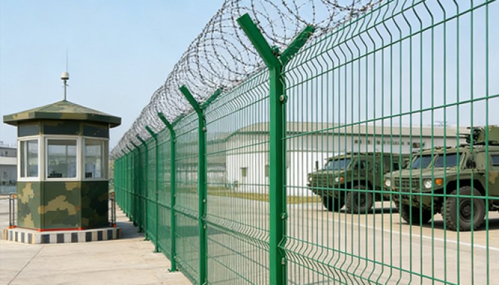 Green high-security metal fence topped with razor wire surrounding a secured facility with guard post